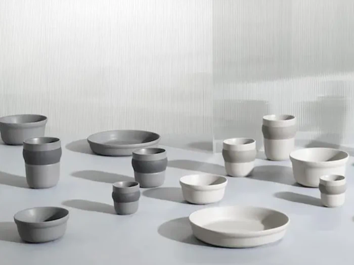 En samling af minimalistiske keramiske skåle og kopper i neutrale grå og hvide toner, arrangeret på en glat overflade med blød belysning og en struktureret lys baggrund.