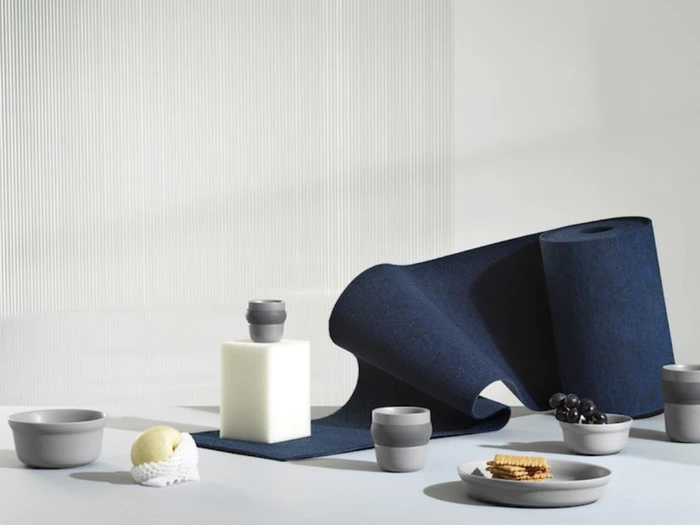 Et minimalistisk stilleben med grå skåle, kopper, tallerkener, en citron, vindruer, kiks og et mørkeblåt struktureret stof, der er rullet ud over en hvid overflade og baggrund.