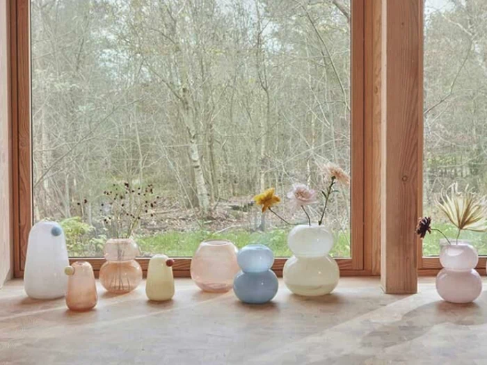 En række pastelfarvede glasvaser med tørrede blomster står i en vindueskarm af træ med udsigt til træer og græs udenfor.