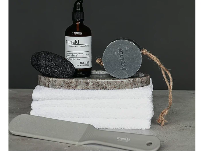 Hudplejeartikler, herunder en pimpsten, kropsolie, sæbe på et reb, stablede hvide håndklæder og en fodfil, alt sammen udstillet mod en mørk baggrund.