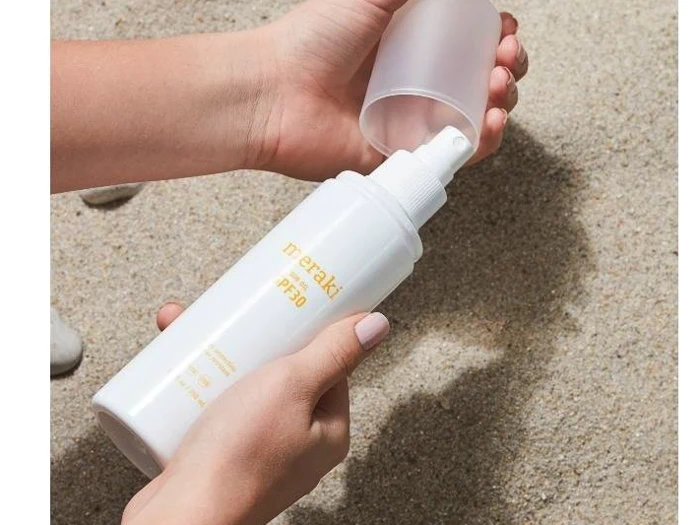 En person holder en hvid flaske Meraki SPF 30 solcreme og tager hætten af, mens der er sand i baggrunden.