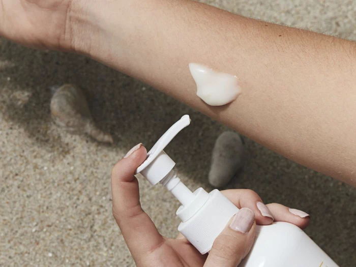 En person pumper hvid lotion på sin underarm, mens han holder en flaske, med sand og muslingeskaller i baggrunden.