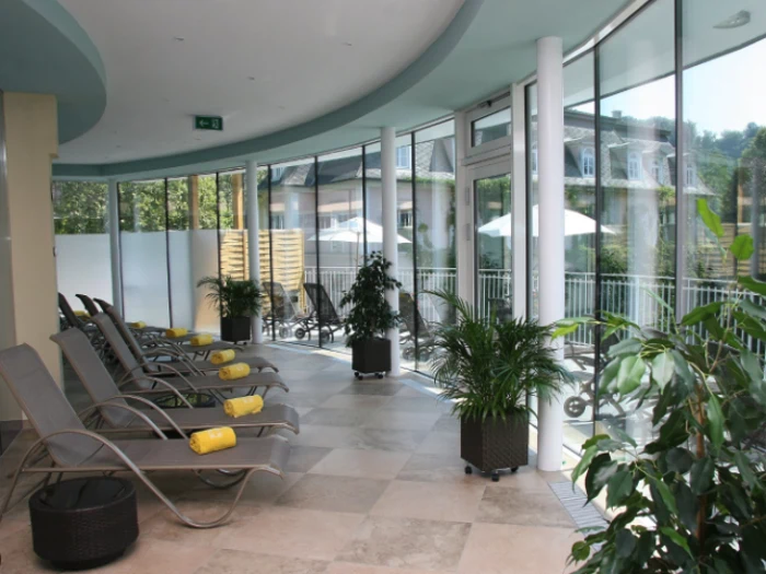 Solbeskinnet indendørs lounge med liggestole, gule håndklæder og potteplanter ved store glasvinduer med udsigt til en balkon og grønne områder.