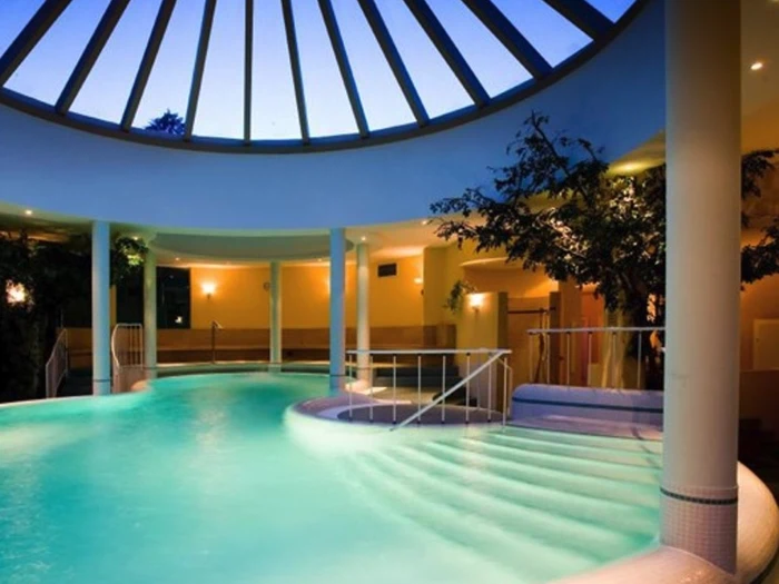 Indendørs pool med glødende turkis vand, omgivet af søjler og træer, under en glaskuppel, der lukker aftenlyset ind. Hvide trin fører ned i vandet.