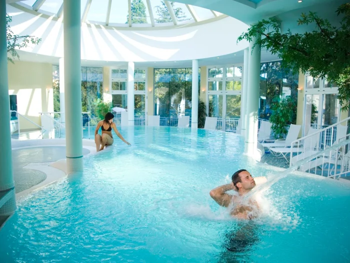 En mand nyder en vandstrålemassage i en indendørs pool, mens en kvinde går ned ad en trappe i vandet under en lys glaskuppel med træer og liggestole i nærheden.