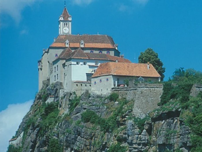 Et historisk slot med røde tage og et klokketårn ligger på toppen af en klippe under en blå himmel.
