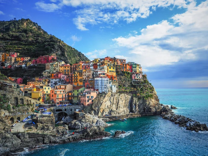 Farverige klippehuse i Manarola, Cinque Terre, Italien, med udsigt over det blå Middelhav med klippekyst og frodige bjergskråninger under en delvis overskyet himmel.