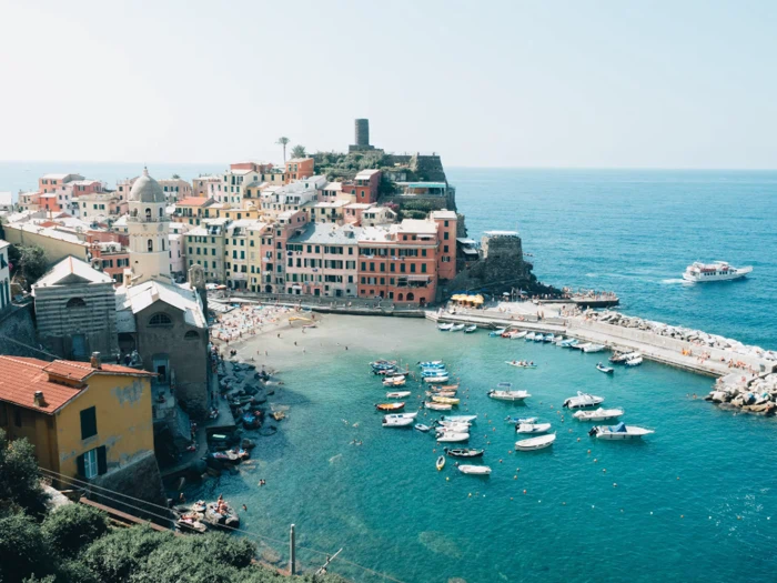 Farverige bygninger ligger langs en kystby med et borglignende tårn, små både i en turkisblå havn og folk, der slapper af på stranden under en klar blå himmel.