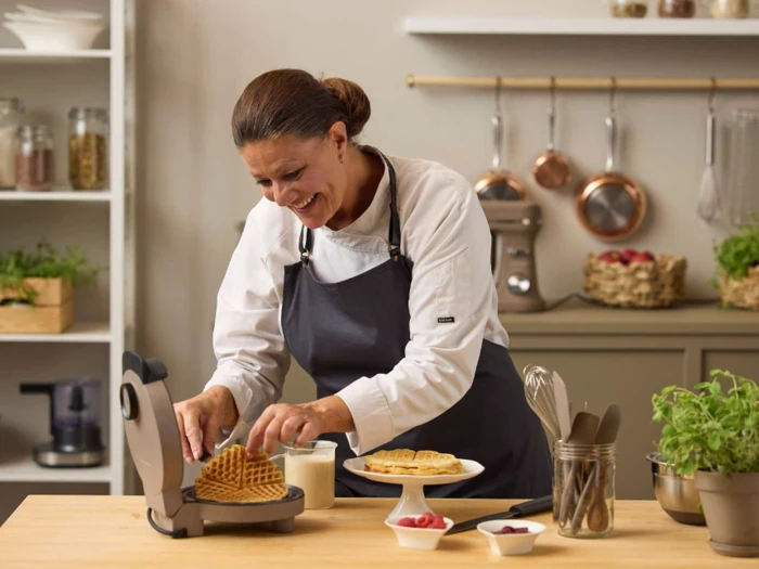 En smilende kvinde i kokkeuniform laver vafler i et køkken og placerer friskbagte vafler på en tallerken omgivet af frugtskåle og køkkenredskaber.
