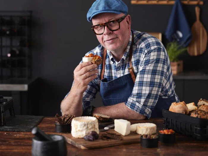 En smilende mand med briller og blå kasket, iført ternet skjorte og forklæde, holder ost og brød ved et rustikt bord med forskellige oste og snacks.