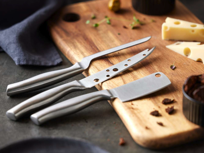 Tre osteknive af metal ligger på et skærebræt af træ med skiver af ost, krydderurter og små stykker af anden mad i nærheden.