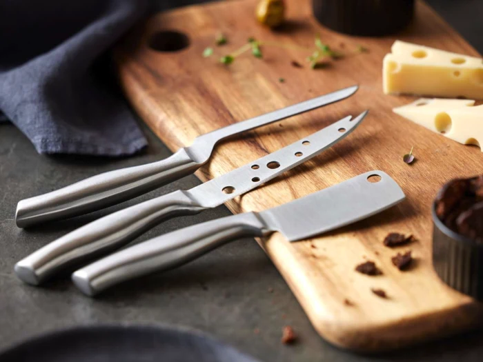 Tre osteknive af metal ligger på et skærebræt af træ med skiver af ost, krydderurter og små stykker af anden mad i nærheden.