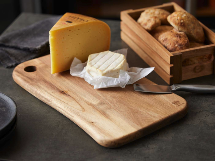 Et træbræt med to slags ost og en kniv ved siden af en trækasse fyldt med rundstykker på en mørk bordplade.
