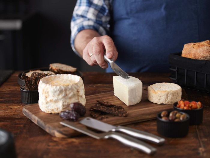 En person skærer en kile af blød ost på et træbræt med forskellige oste, brød, oliven og tørret frugt på et rustikt bord.