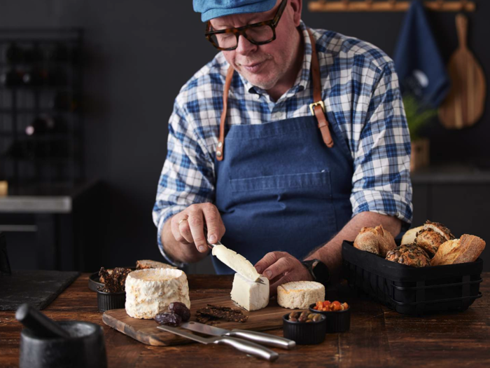 En mand i blåt forklæde og kasket skærer ost ved et træbord med forskelligt brød, dadler og små skåle med mad.
