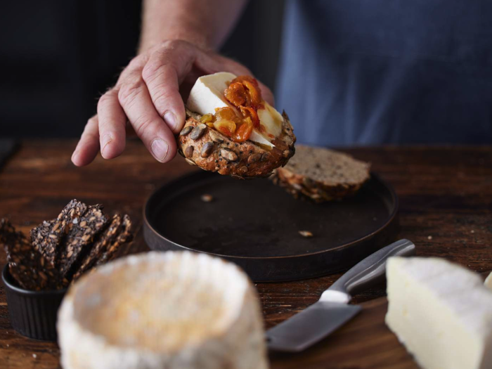 En hånd holder en skive brød med kerner, ost og madpynt over en mørk tallerken med ostekiler og kiks på et træbord.