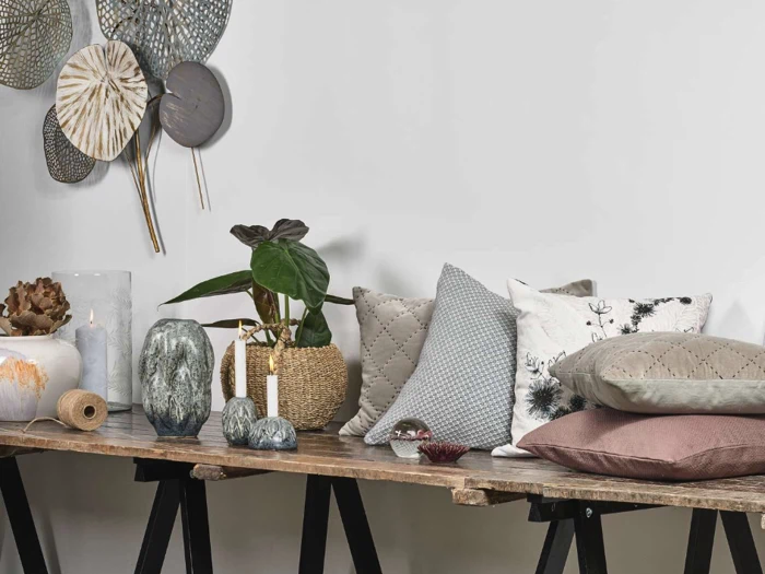 Et rustikt træbord med dekorative puder, stearinlys, potteplanter, en rulle sejlgarn og væghængt bladkunst mod en hvid baggrund.