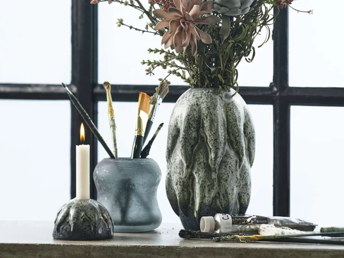 Keramikvaser med blomster, pensler og et tændt stearinlys står på en flade foran et vindue med sorte rammer. Farvetuber og en paletkniv står i nærheden.