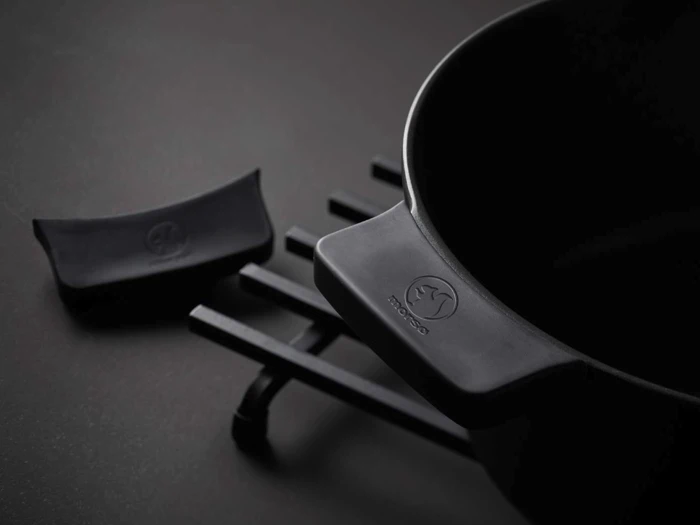 Et nærbillede af en sort støbejernsgryde med håndtag, der hviler på en bordskåner, og et matchende sort silikonehåndtag i nærheden på en mørk overflade.