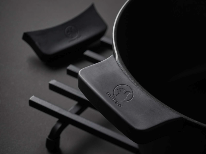 Nærbillede af en sort støbejernsgryde på en sort bordskåner med et matchende sort låghåndtag ved siden af. Grydehåndtaget har et præget logo.