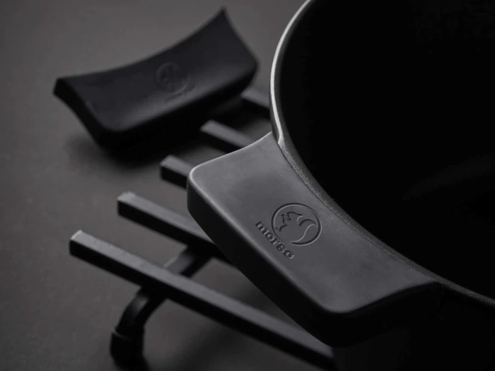 Nærbillede af en sort støbejernsgryde på en sort bordskåner med et matchende sort låghåndtag ved siden af. Grydehåndtaget har et præget logo.