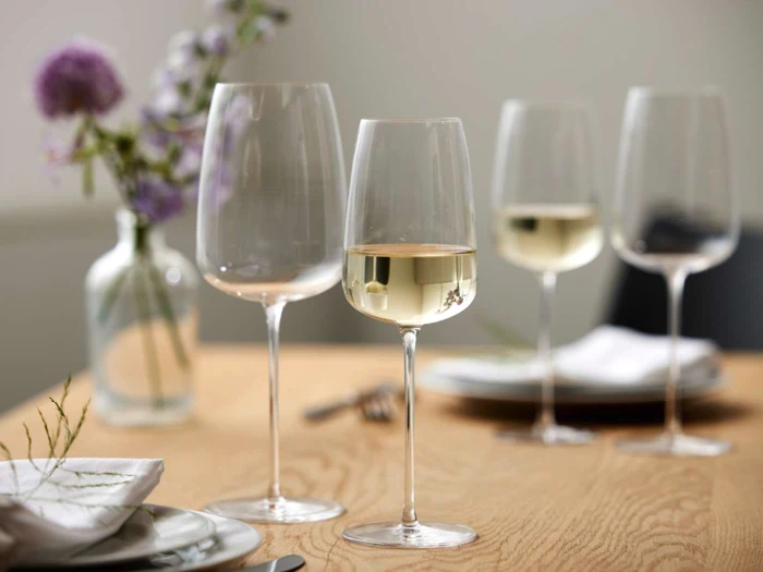 Fire vinglas, to fyldt med hvidvin, er anbragt på et træbord med tallerkener, servietter og en vase med lilla blomster i baggrunden.