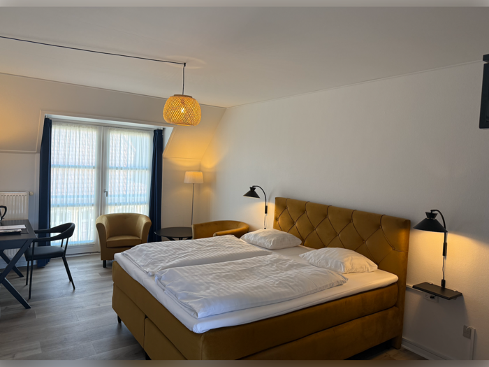 Et lyst hotelværelse med en stor seng, sennepsgult hovedgærde og stole, et skrivebord, lamper og et stort vindue med gennemsigtige gardiner og mørkeblå forhæng.