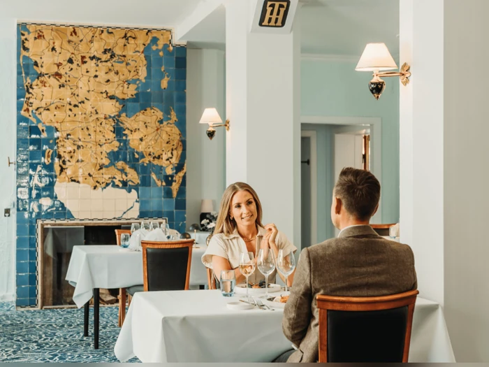 En kvinde og en mand sidder ved et veldækket bord på en restaurant og taler og smiler. Bag dem kan man se blå og gyldne fliser på væggen.