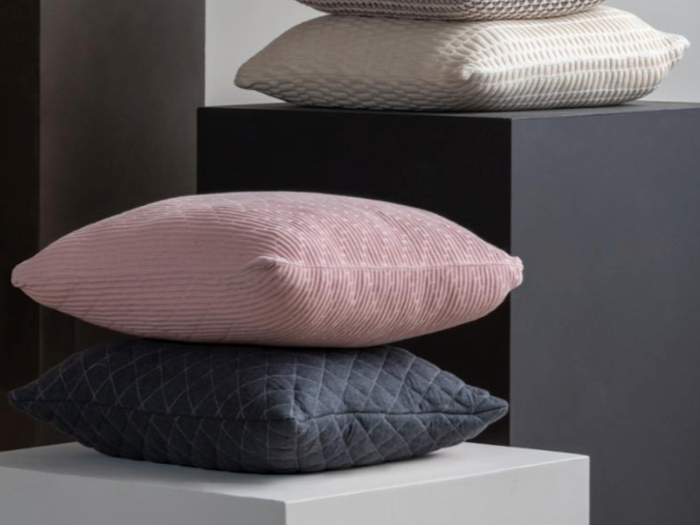 Fire teksturerede puder i pastelfarver og neutrale toner er stablet på moderne firkantede piedestaler mod en ensfarvet baggrund.