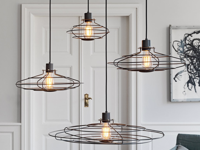 Fire synlige glødetrådspendler med wireframe-skærme hænger fra loftet foran en hvid dobbeltdør i et moderne, minimalistisk opholdsrum.