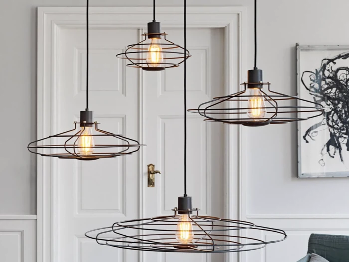 Fire synlige glødetrådspendler med wireframe-skærme hænger fra loftet foran en hvid dobbeltdør i et moderne, minimalistisk opholdsrum.