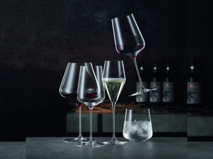 Fem elegante glas - tre med rødvin, et med mousserende vin og et med isvand - står på et bord; et rødvinsglas står på skrå og balancerer på et andet.