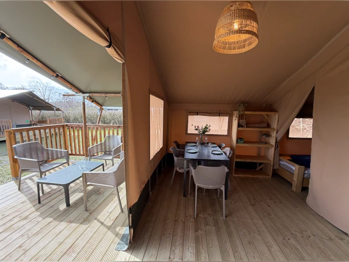 Interiør i et glampingtelt med spisebord, stole, hylder, en seng, der er delvis synlig, og en udendørs terrasse med flere siddepladser og et bord, alt sammen på trægulv.