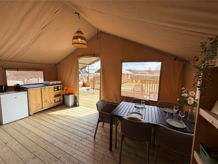 Et hyggeligt glampingtelt med et lille køkken, et spisebord med plads til fire og store vinduer med udsigt til et trædæk og den udendørs natur.