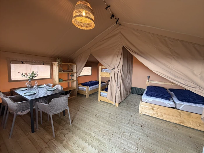Indvendigt billede af et rummeligt glampingtelt med en dobbeltseng, en køjeseng, et spisebord med stole, hylder og dæmpet belysning, alt sammen på et trægulv.