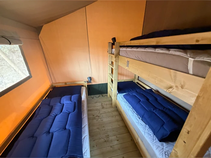 Interiør i et telt med trægulv, en enkeltseng til venstre og en køjeseng af træ med blå tæpper ved siden af et vindue og en lampe.