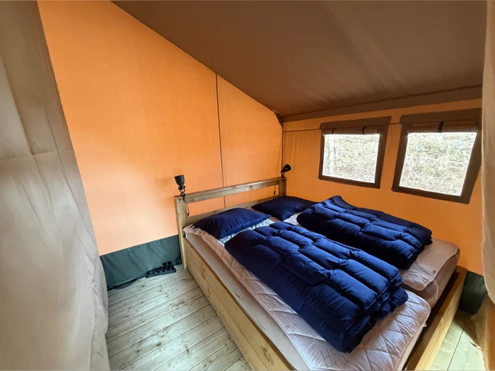 Et hyggeligt teltinteriør med trægulv, dobbeltseng med to blå soveposer og to vinduer, der lukker naturligt lys ind.