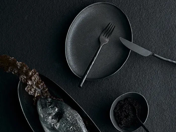 En borddækning med mørkt tema med en fisk på et fad, en struktureret tallerken med en gaffel, en kniv og en lille skål med kaviar, alt sammen på en sort overflade.