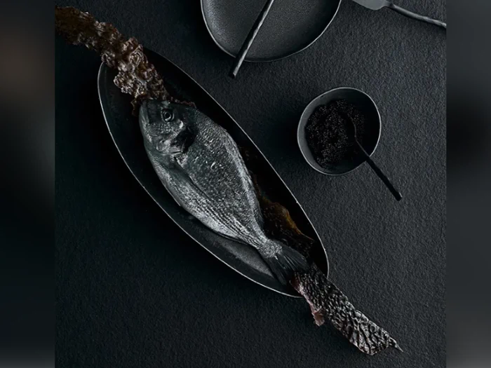 En hel fisk på en mørk oval tallerken, serveret med et stykke sprødt fiskeskind. Sorte skåle og redskaber er arrangeret på en mørk struktureret overflade.