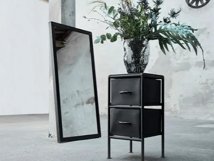 Et natbord i sort metal med to skuffer og en vase med grønne grene står ved siden af et højt, fritstående spejl i et minimalistisk rum.