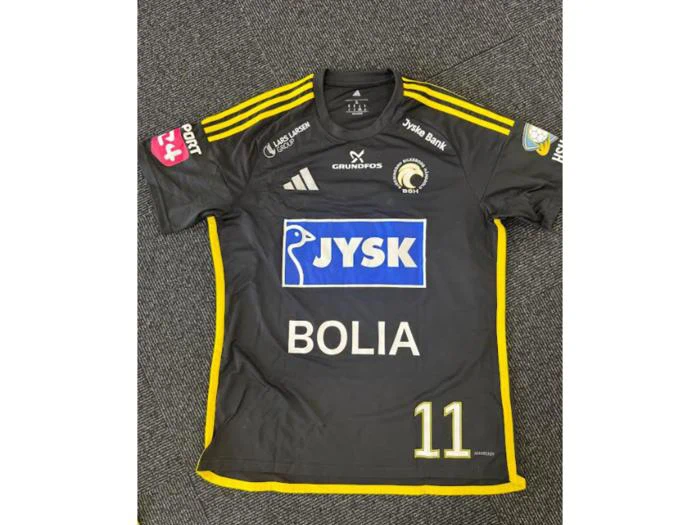 En sort og gul sportstrøje med sponsorlogoer, nummer 11 og et cirkulært emblem på brystet, der lå fladt på et mørkt tæppe.