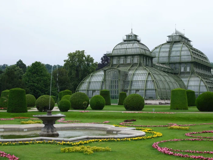 Et stort glasdrivhus med kuplede tage står i en velplejet have med trimmede buske, et springvand og farverige blomsterbede under en overskyet himmel.