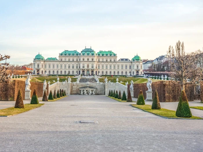 Barokpalads med grønt tag, symmetriske haver, klippede hække, skulpturer og et springvand foran, set fra en bred sti under en klar himmel.