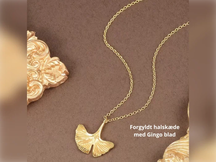 En guldbelagt halskæde med et vedhæng af ginkgoblade vises på en brun baggrund med udsmykket gulddekor. Den danske tekst lyder: Forgyldt halskæde med Gingo blad.