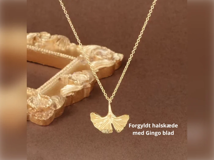 En guldbelagt halskæde med et vedhæng af ginkgoblade ses ved siden af en udsmykket guldramme. Den danske tekst lyder "Forgyldt halskæde med Gingo blad.".