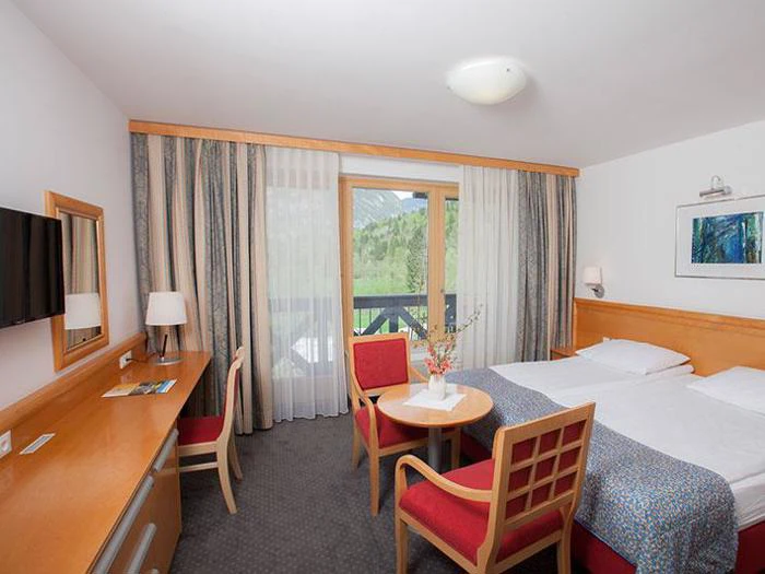 Et hotelværelse med dobbeltseng, skrivebord, tv, to røde stole, et rundt bord og et vindue med bjergudsigt og gennemsigtige gardiner.