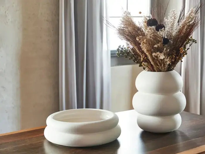 En hvid, afrundet vase med tørrede blomster står ved siden af en tilsvarende tom skål på et træbord med lyse gardiner i baggrunden.