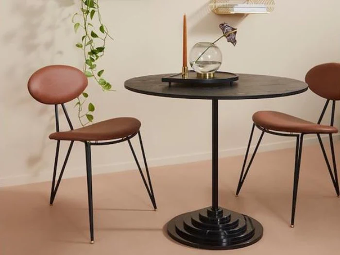 Et moderne rundt sort bord med et guldlys, en glasskål og to brune stole med metalben; en hængende plante i baggrunden.