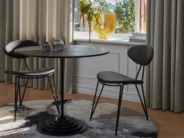 Et rundt sort bord med to sorte stole står på et mønstret tæppe nær et vindue med beige gardiner og en glasvase med blomster i vindueskarmen.