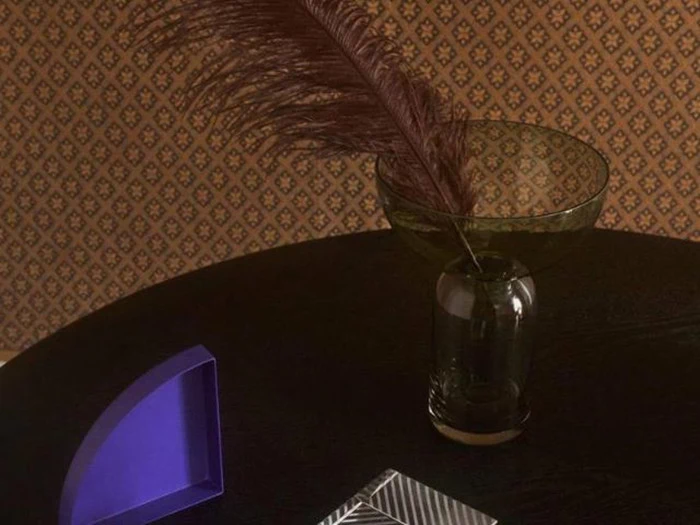 En brun fjer i en glasvase står på et mørkt rundt bord med en lilla geometrisk genstand og en stribet notesbog på en mønstret tapetbaggrund.